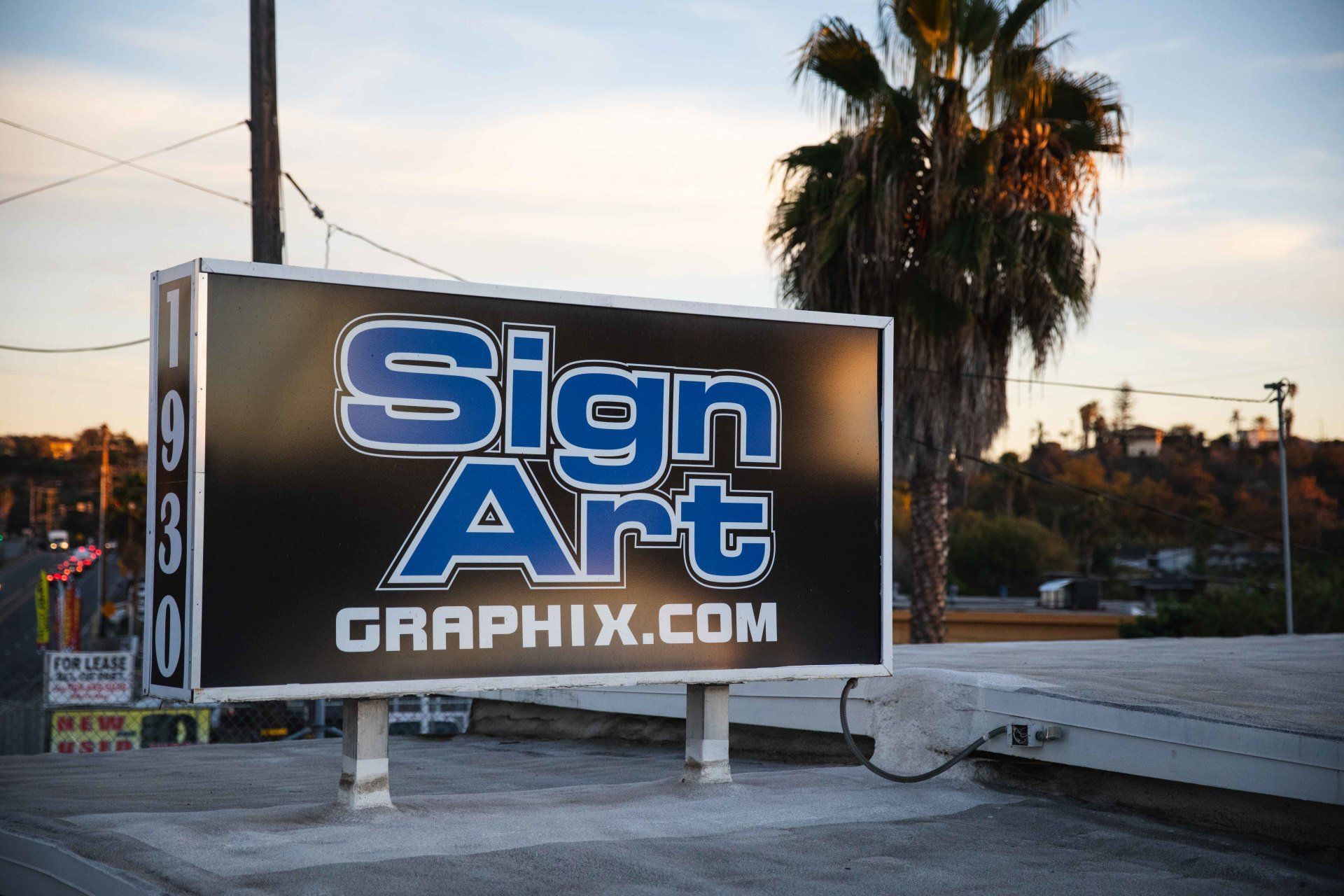 Sign Art Graphix sign in front of a palm tree and cityscape, at dusk.