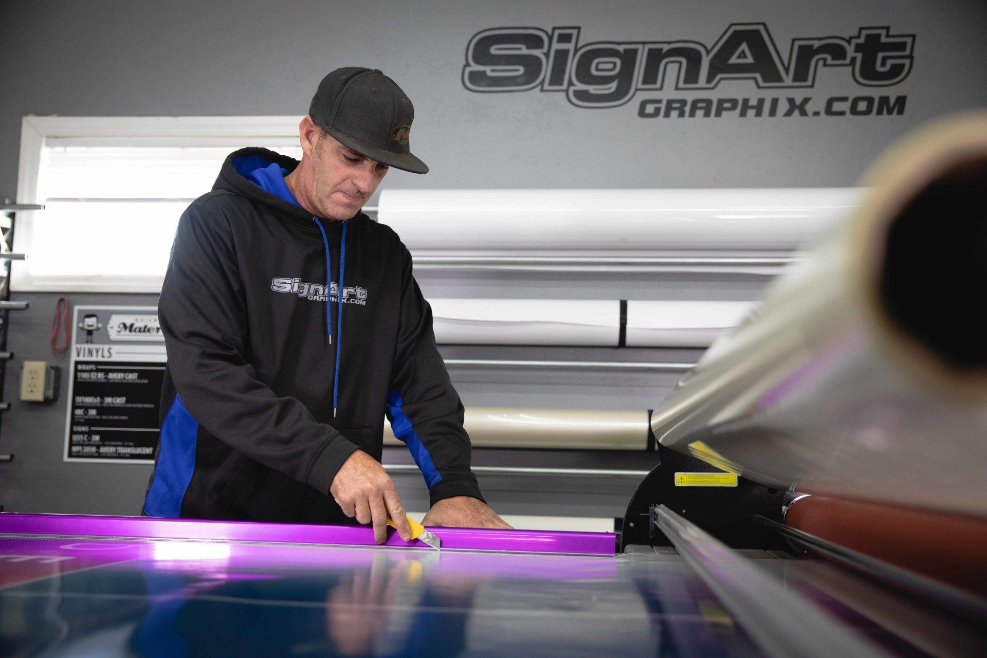 Person operating a large-format printer in a print shop, handling purple vinyl film under bright lights