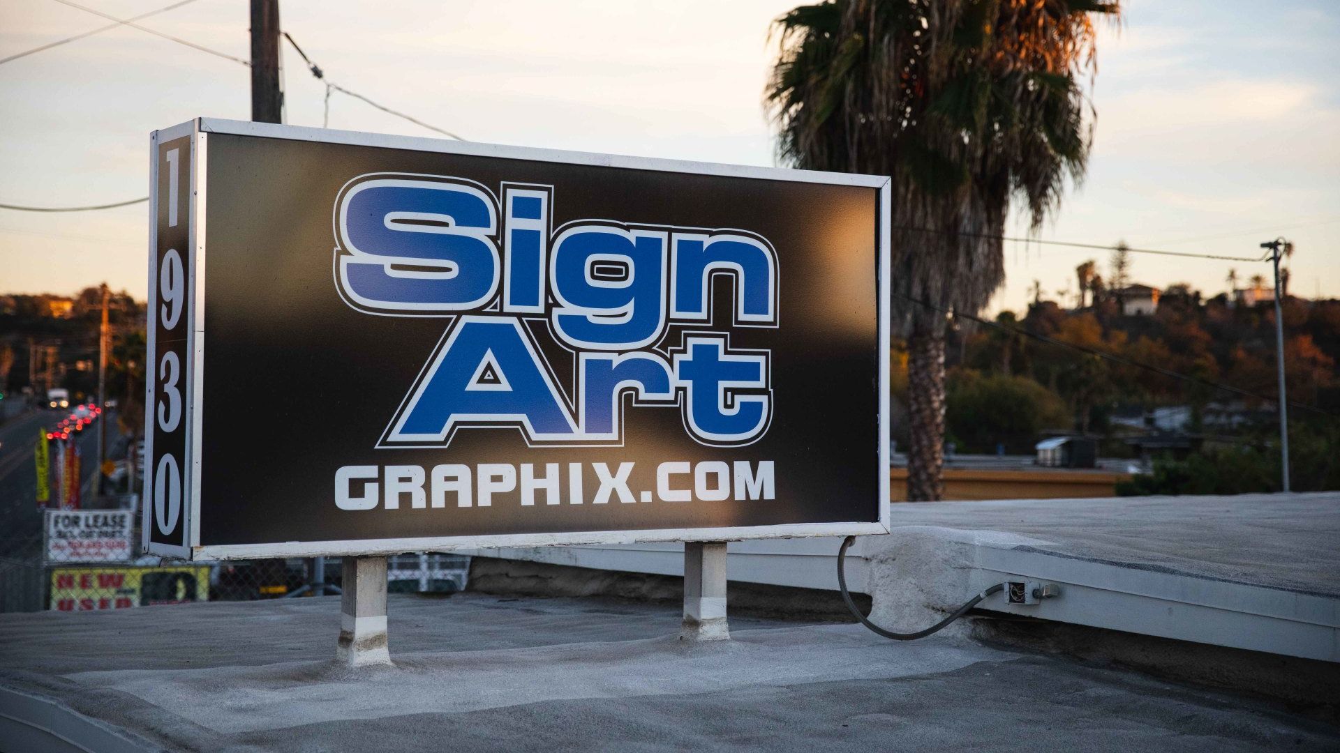 Sign Art Graphix sign in front of a palm tree, with a street and sunset visible.