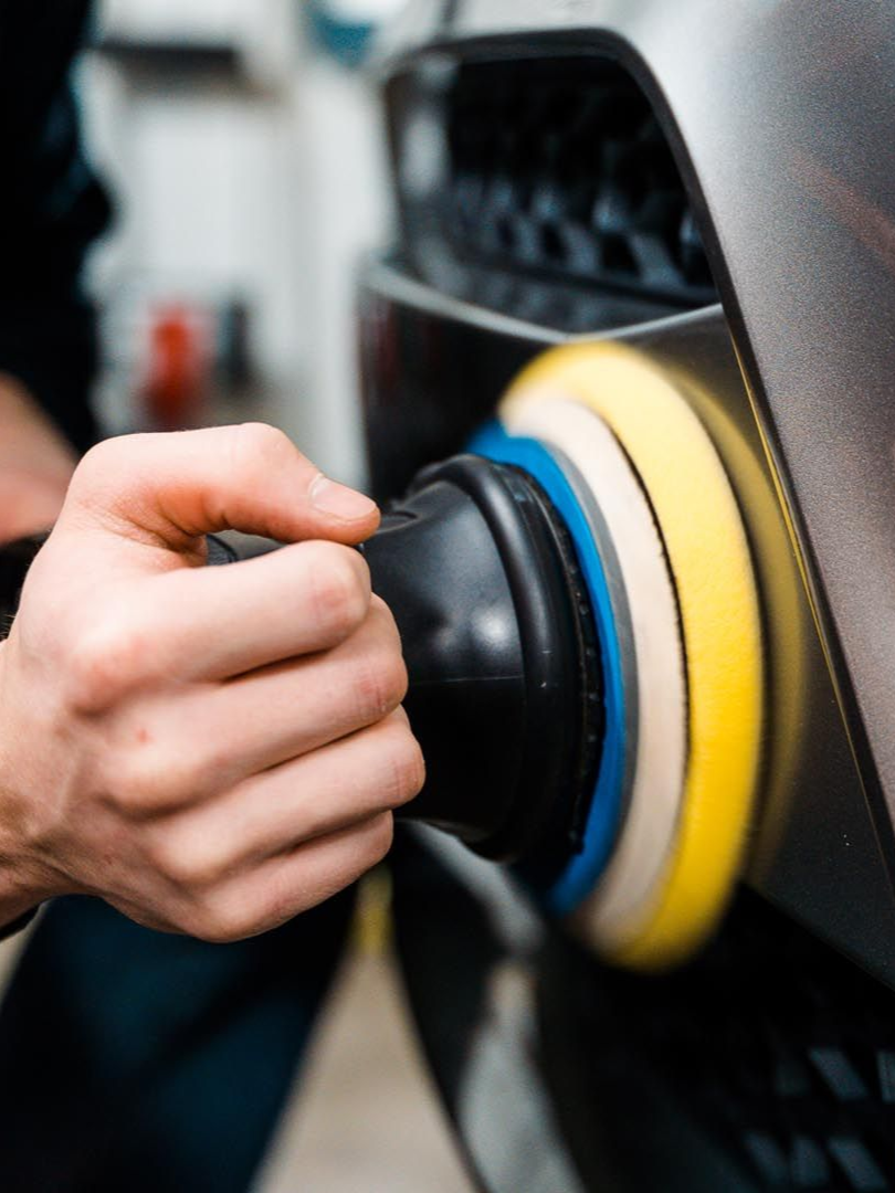 Person polishes a gray car bumper with a rotary buffer using multi-colored polishing pads.