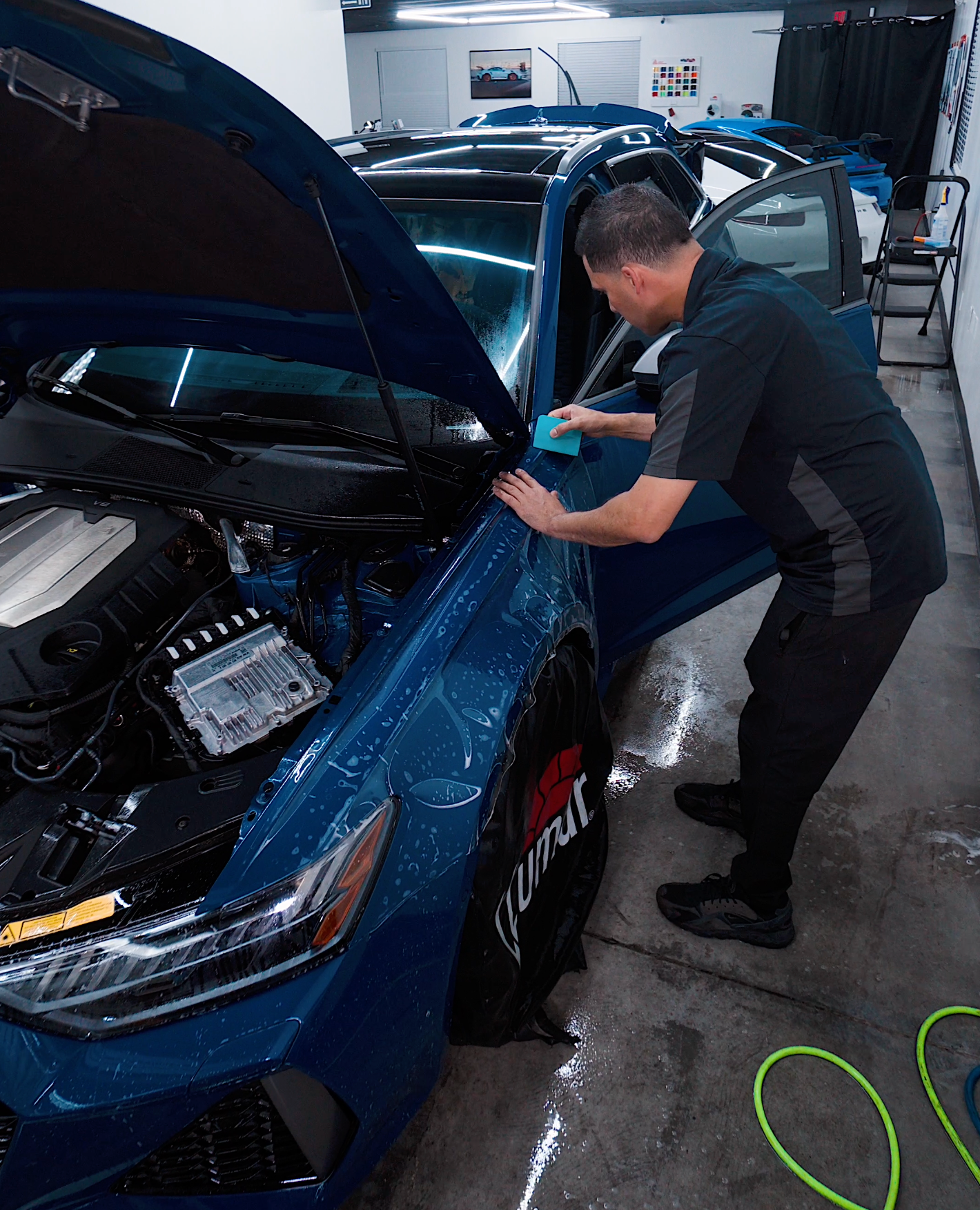 Man applying protective film to a blue car in a workshop.