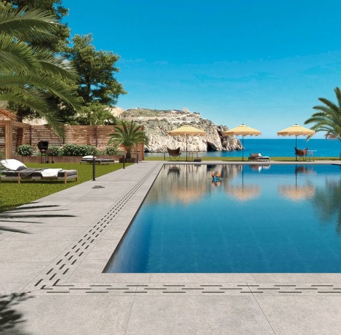 Piscina a sfioro di lusso accanto a palme e lettini, con vista su una costa rocciosa sotto un cielo azzurro e terso
