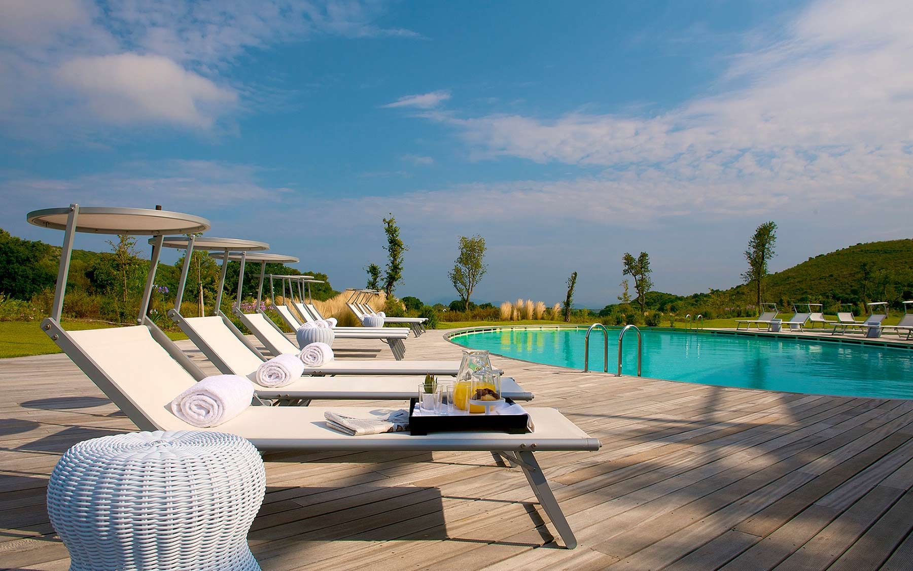 Lettini a bordo piscina accanto a una piscina turchese sotto un cielo azzurro, con un tavolino in vimini in primo piano.