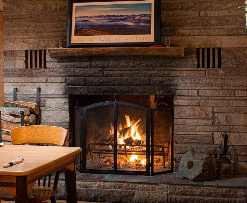 Cozy stone fireplace with burning fire, wood mantel, and framed landscape above a chair and table