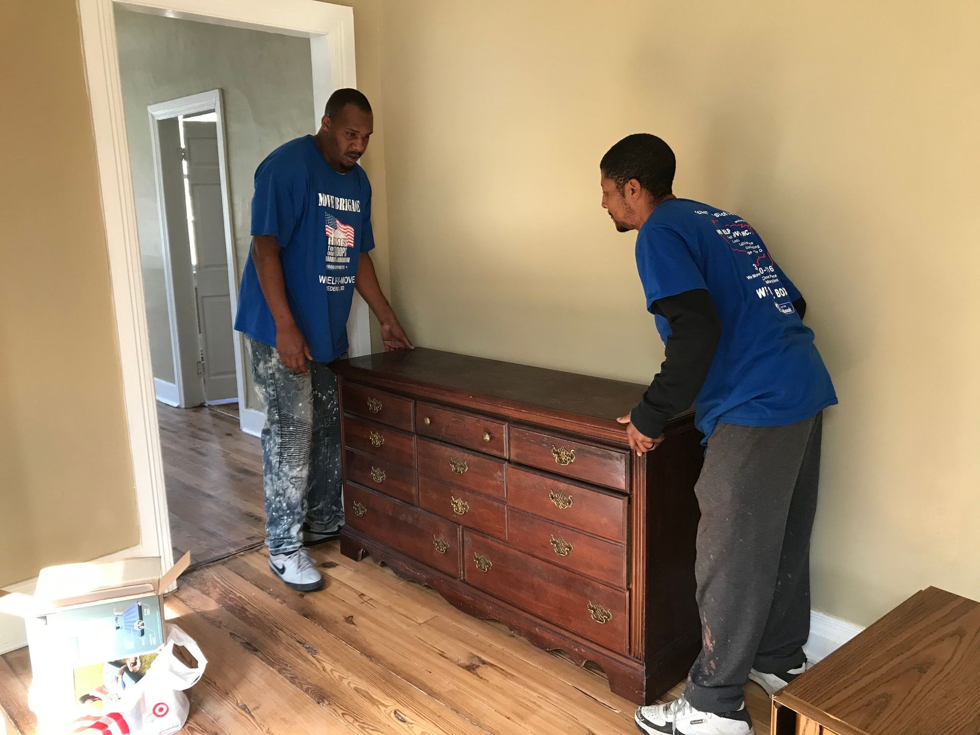 Two movers in blue shirts lift a wooden dresser in a room with light-colored walls.