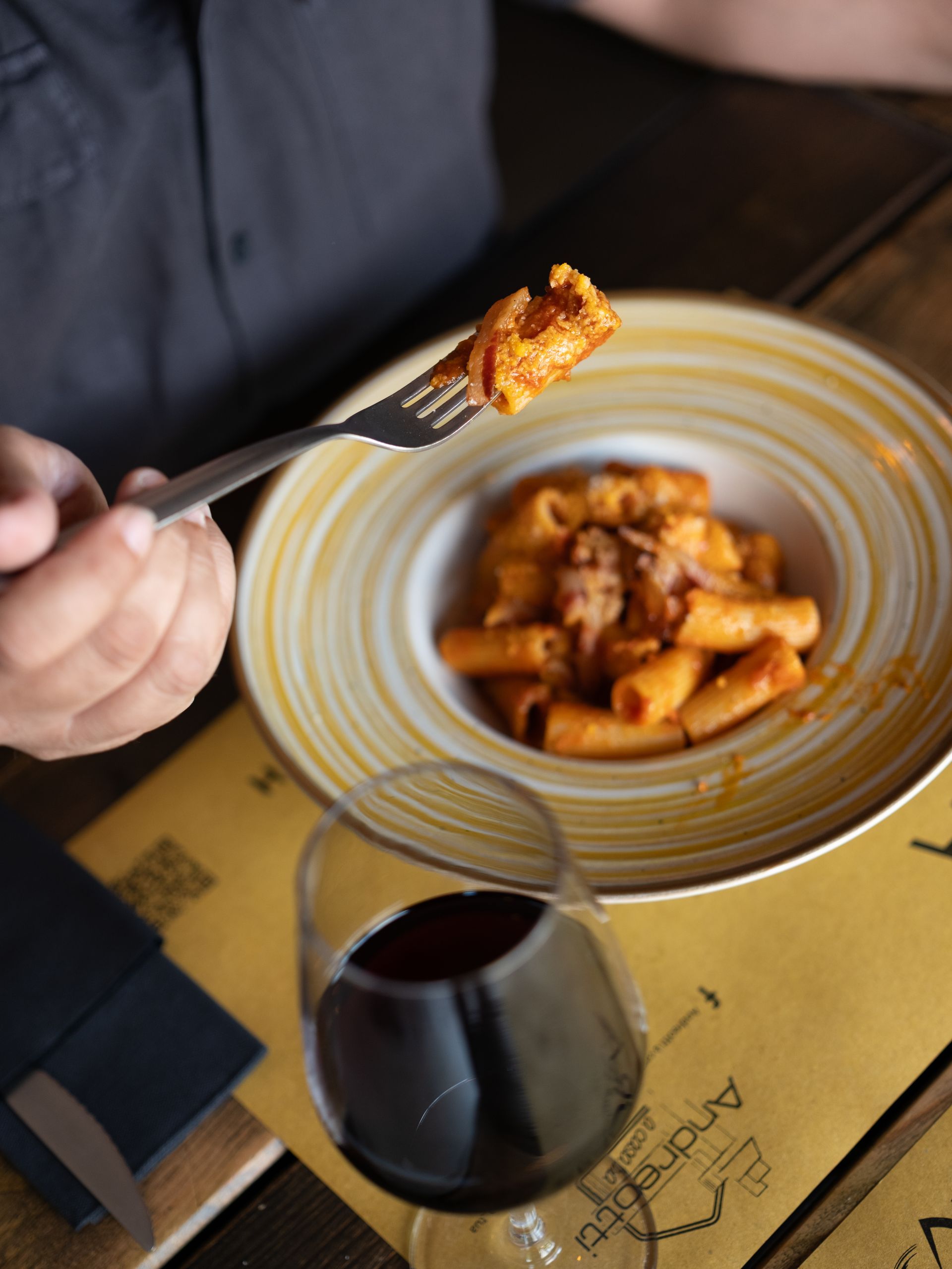Persona che tiene in mano una forchetta con la pasta. Piatto di pasta con sugo rosso, bicchiere di vino rosso, su un tavolo di legno.
