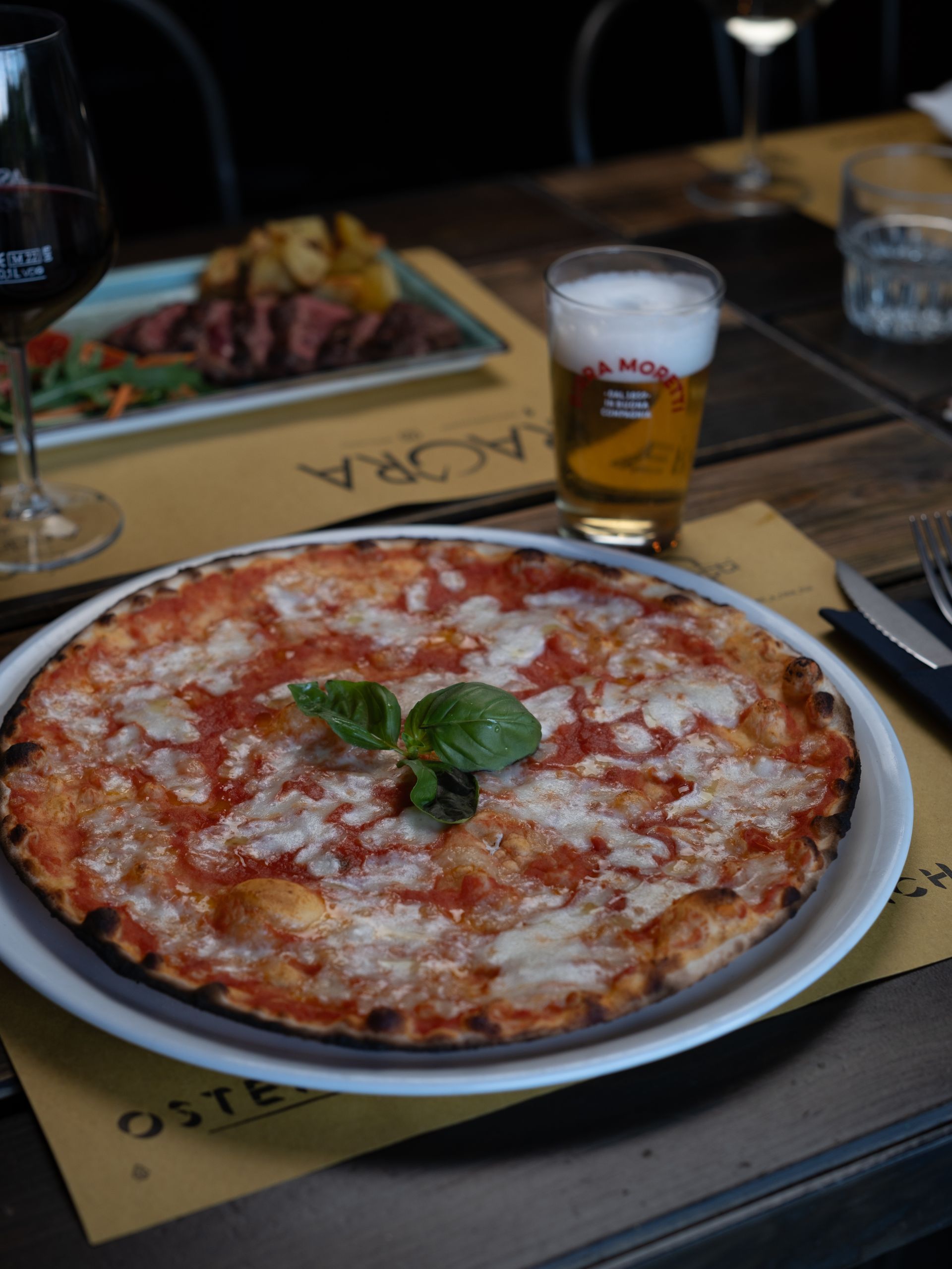 Pizza su un piatto, guarnizione di basilico, con birra, bistecca e vino su un tavolo di legno in un ristorante.