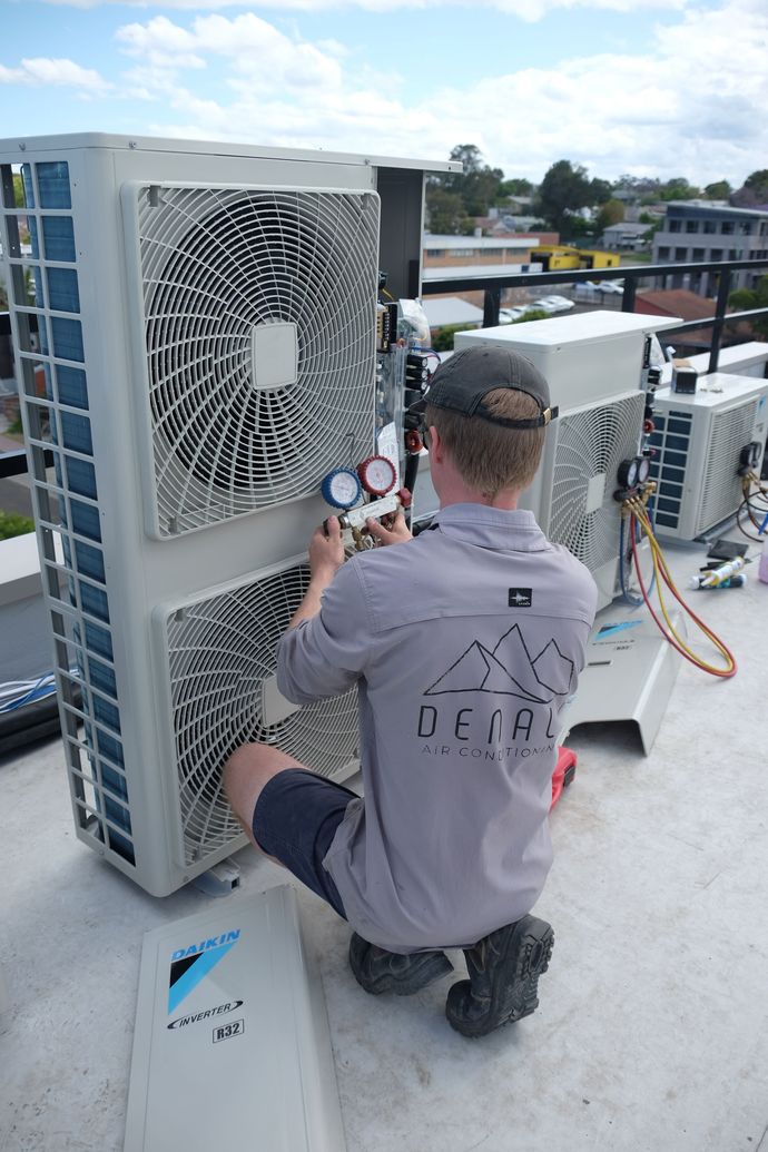 Two Denali Air Conditioning workers in work shirts installing an air conditioner unit next to a building in Nowra