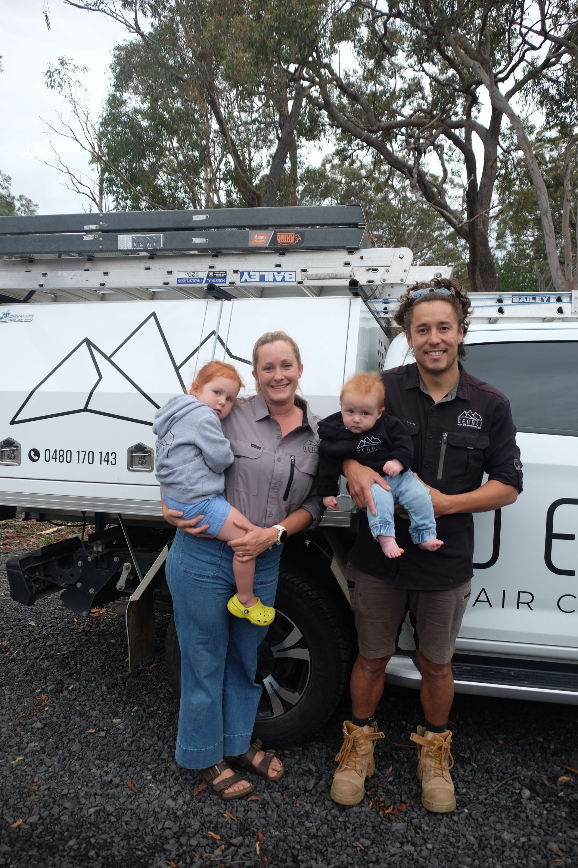 Two Denali Air Conditioning workers in work shirts installing an air conditioner unit next to a building in Nowra