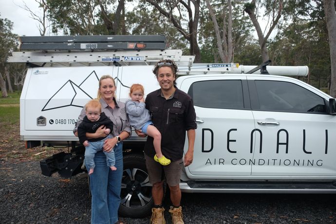 Two Denali Air Conditioning workers in work shirts installing an air conditioner unit next to a building in Nowra