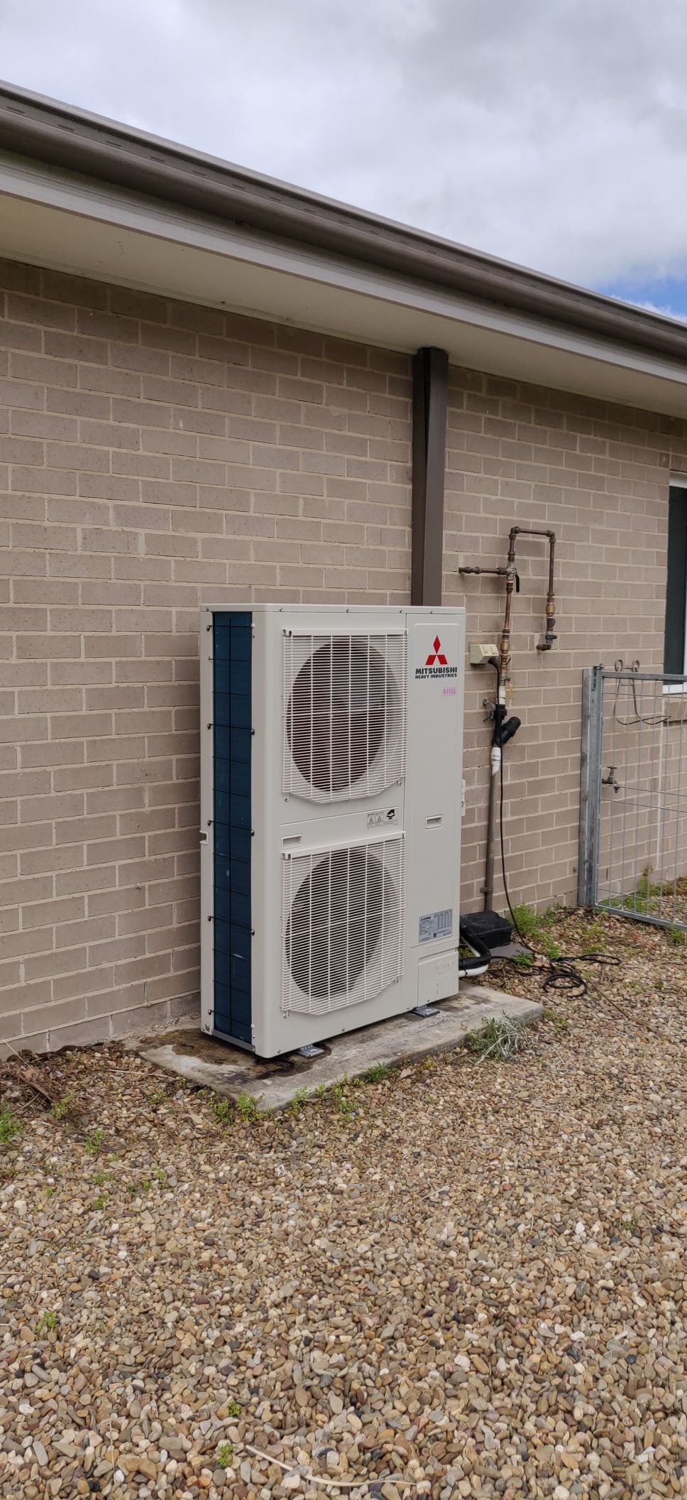 Two Denali Air Conditioning workers in work shirts installing an air conditioner unit next to a building in Nowra