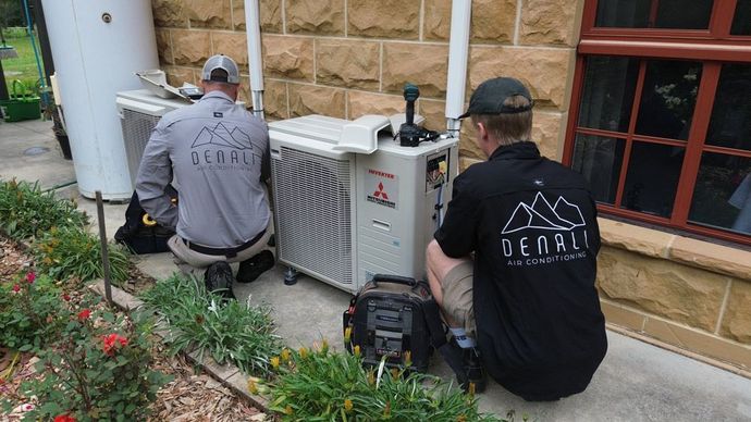 Two Denali Air Conditioning workers in work shirts installing an air conditioner unit next to a building in Nowra