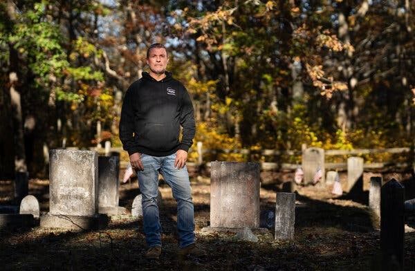 Person standing among old gravestones in a wooded cemetery with autumn leaves.