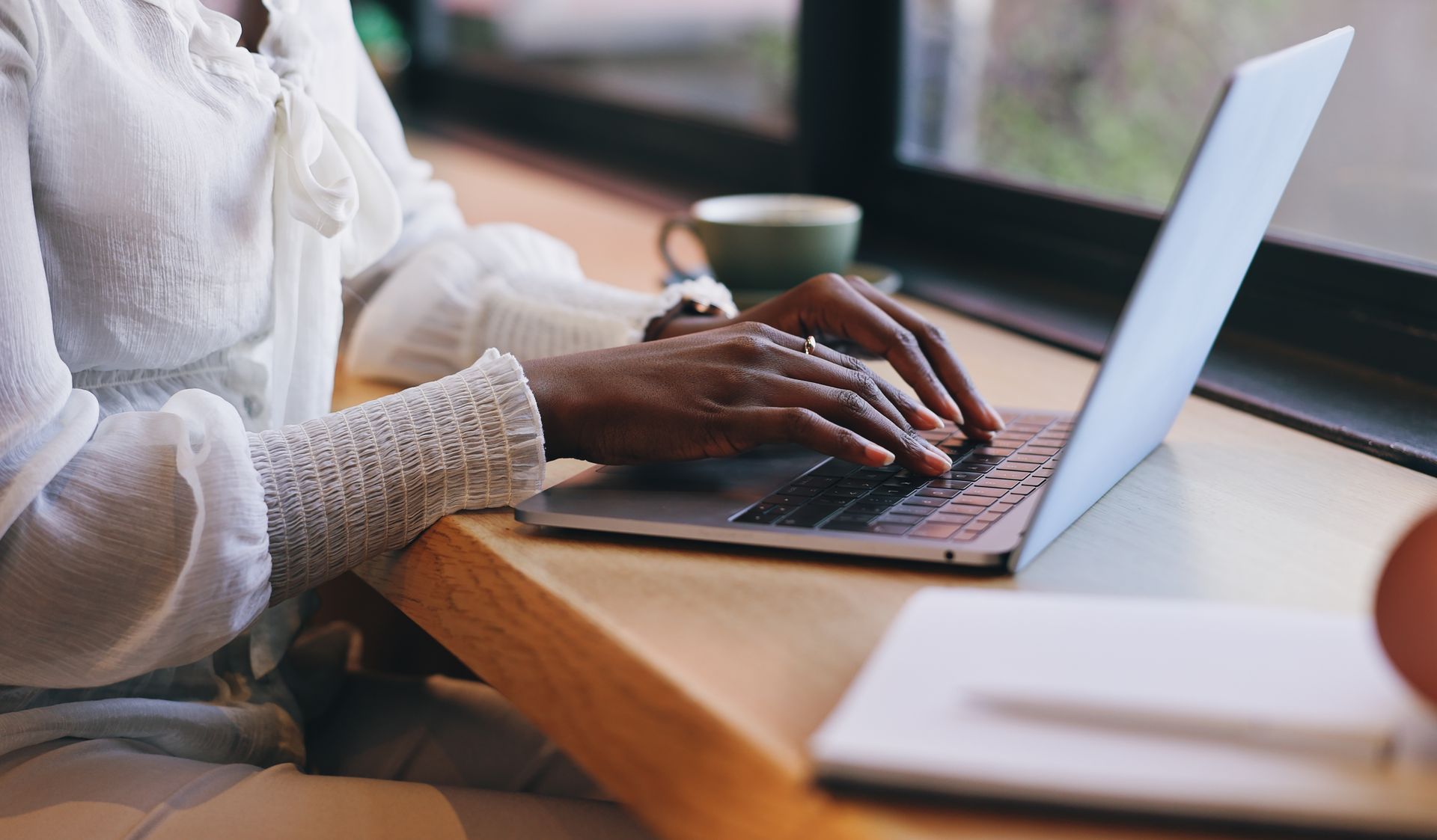 Person typing on a laptop at a wooden table beside a window, with a notebook in the foreground.