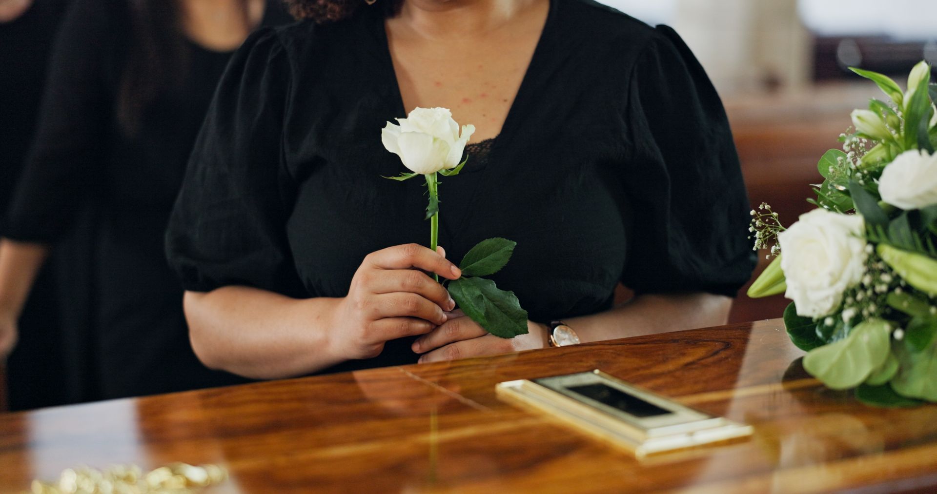 Woman in black dress holding a white rose at a wooden table with a phone and floral arrangement.