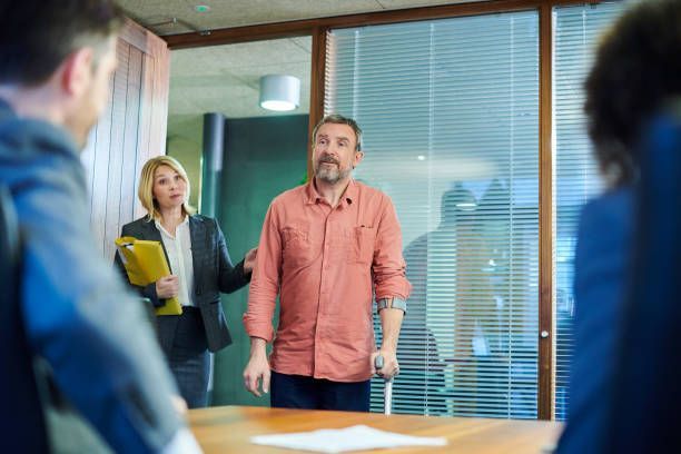 A man with crutches is standing in front of a group of people in a conference room.