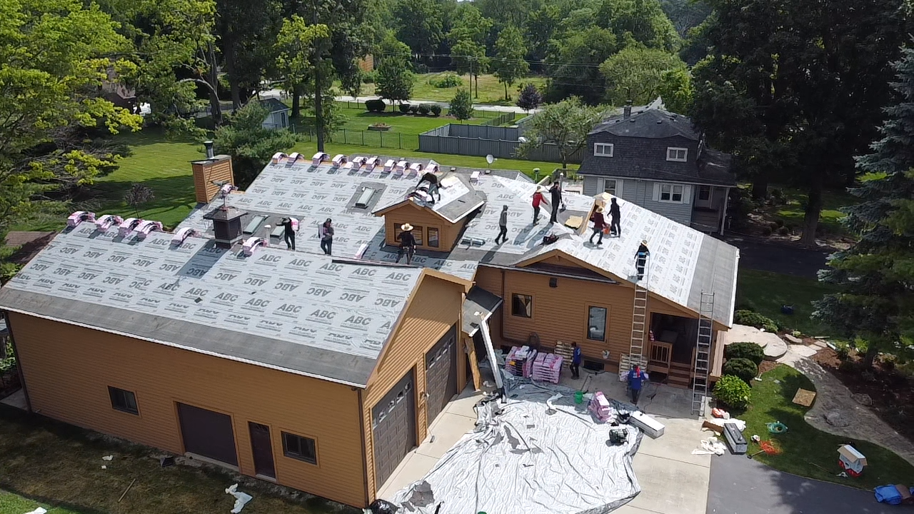 Roofing crew working on a house with gray shingles and white tarps, surrounded by trees and a yard