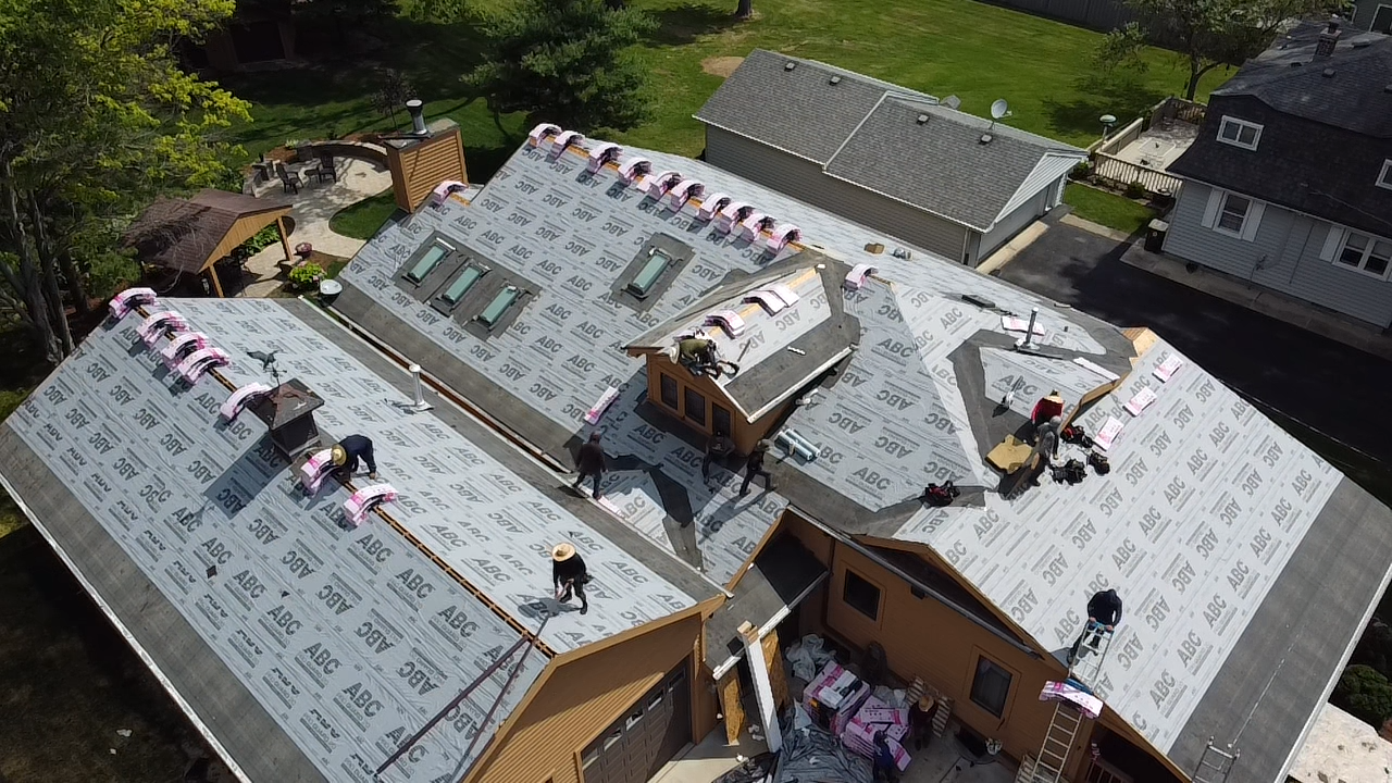 Aerial view of a house roof under repair, with workers and a large white roofing tarp.