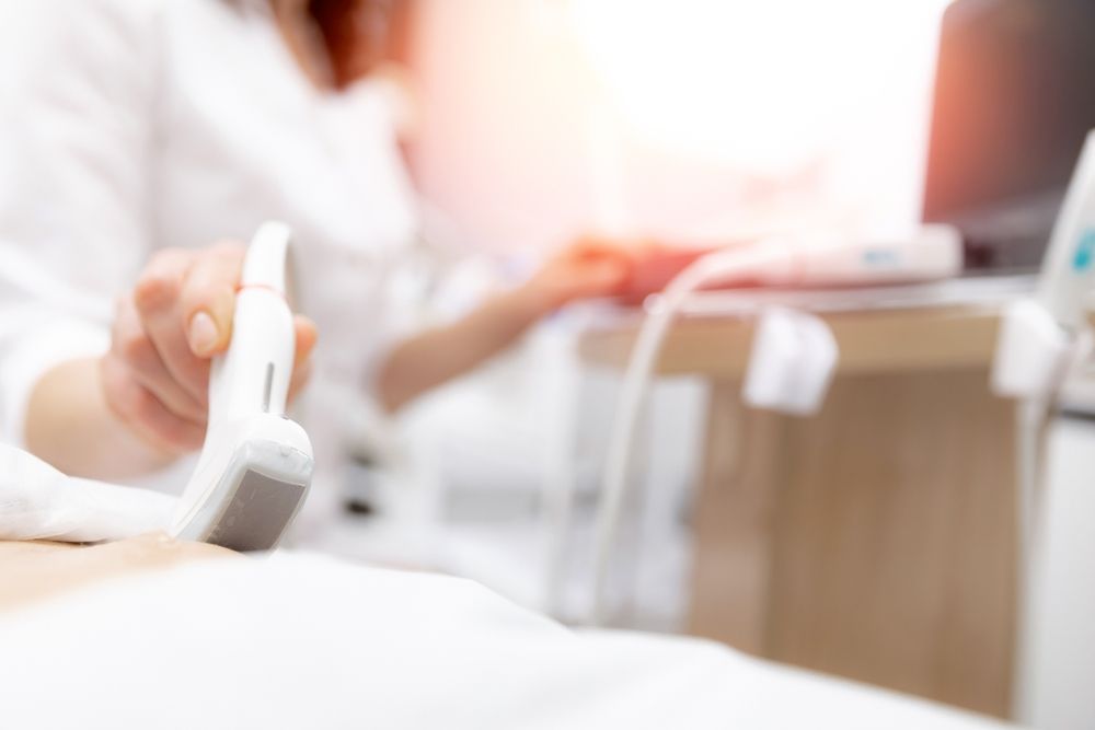 Person performing an ultrasound scan, using a handheld device on a patient's abdomen in a medical setting.