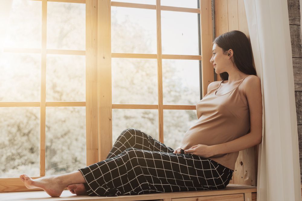 Pregnant woman resting by a sunlit window, leaning back on a cushion and looking outside.