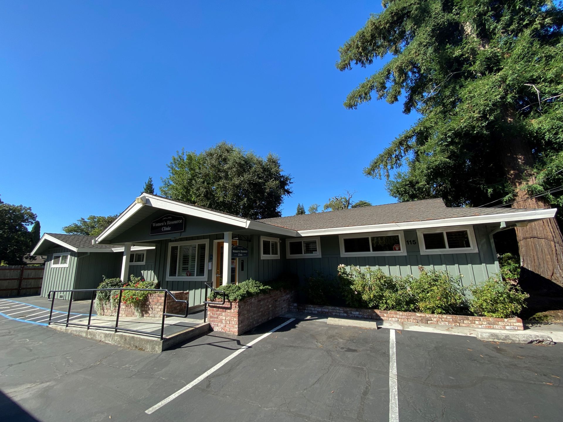 Green building with a sloped roof, surrounded by trees and parking spaces, under a blue sky.