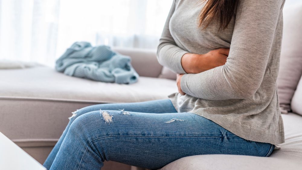 Woman sitting on a couch, clutching her abdomen in pain with a blanket in the background