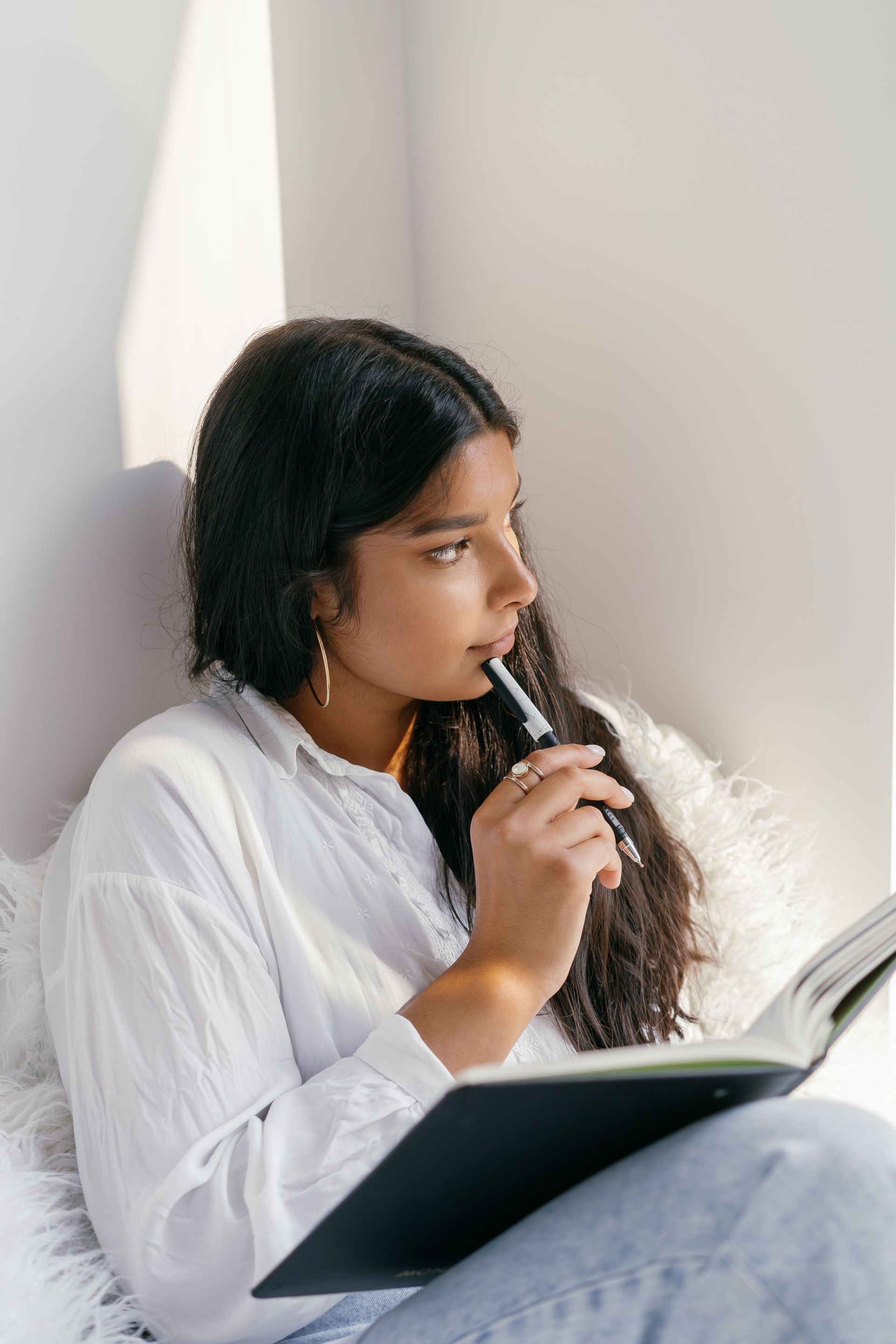 Person sitting by a bright window, reading and holding a pen over an open notebook