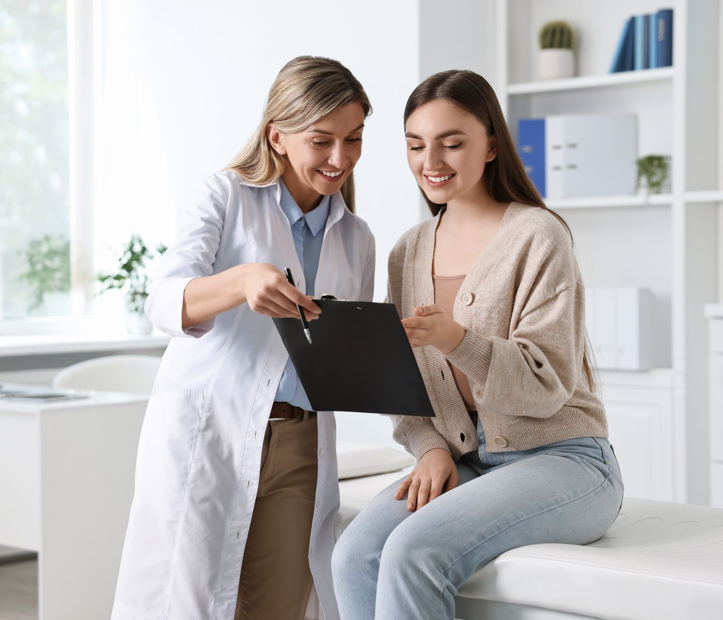 Doctor pointing at a clipboard, smiling at a patient sitting on an examination table in a medical office.