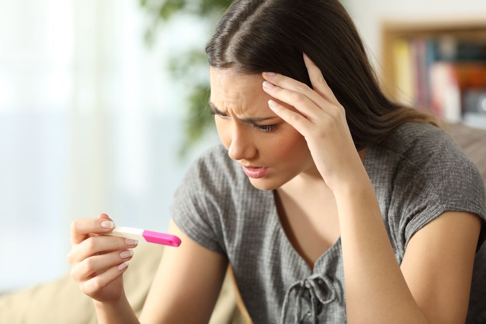 Woman looking worried at a positive pregnancy test, holding her head indoors.