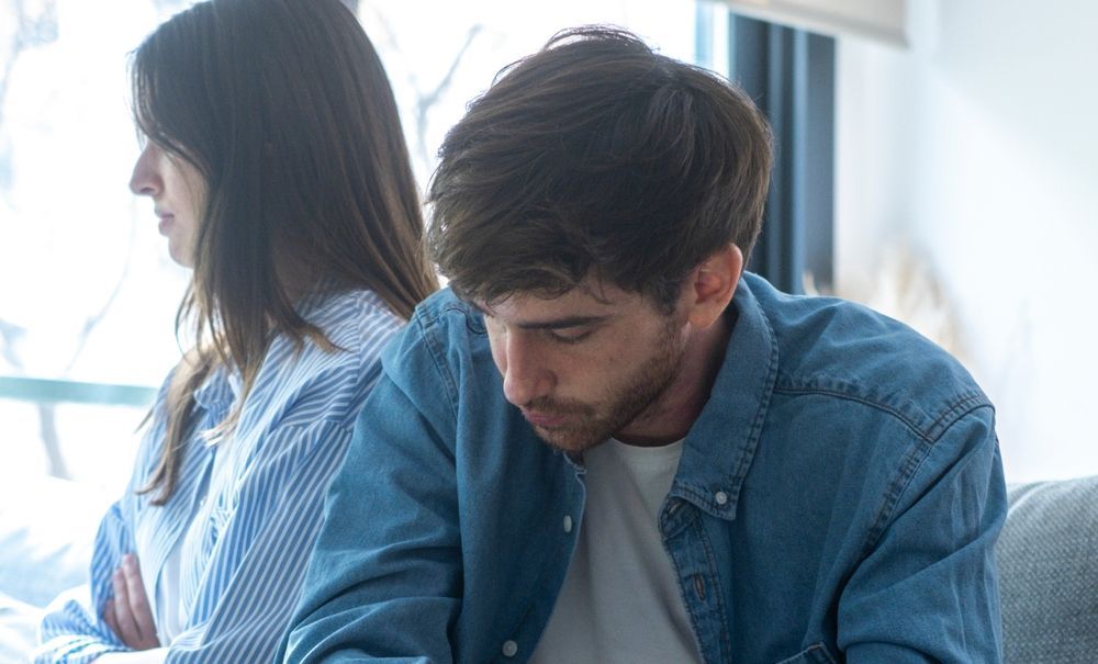 Two people sitting back to back by a bright window, both looking down thoughtfully.