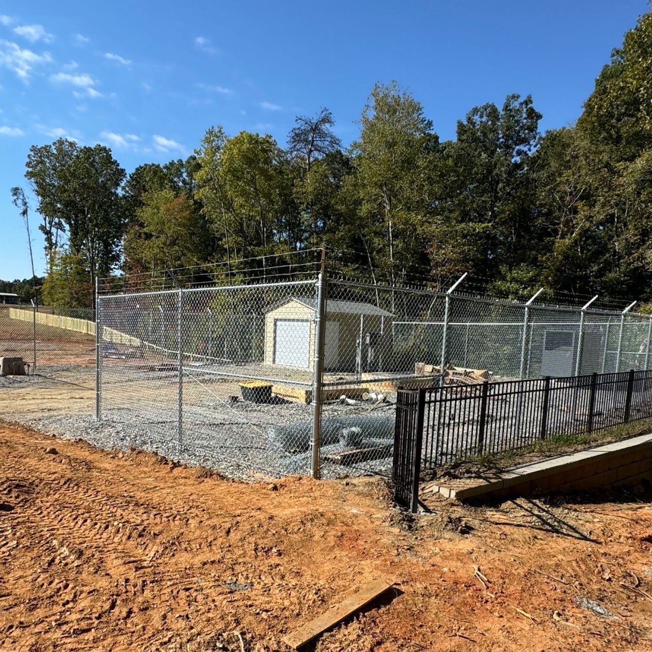 Chain-link fenced utility area with small shed and gravel, beside a dirt road and trees under a blue sky
