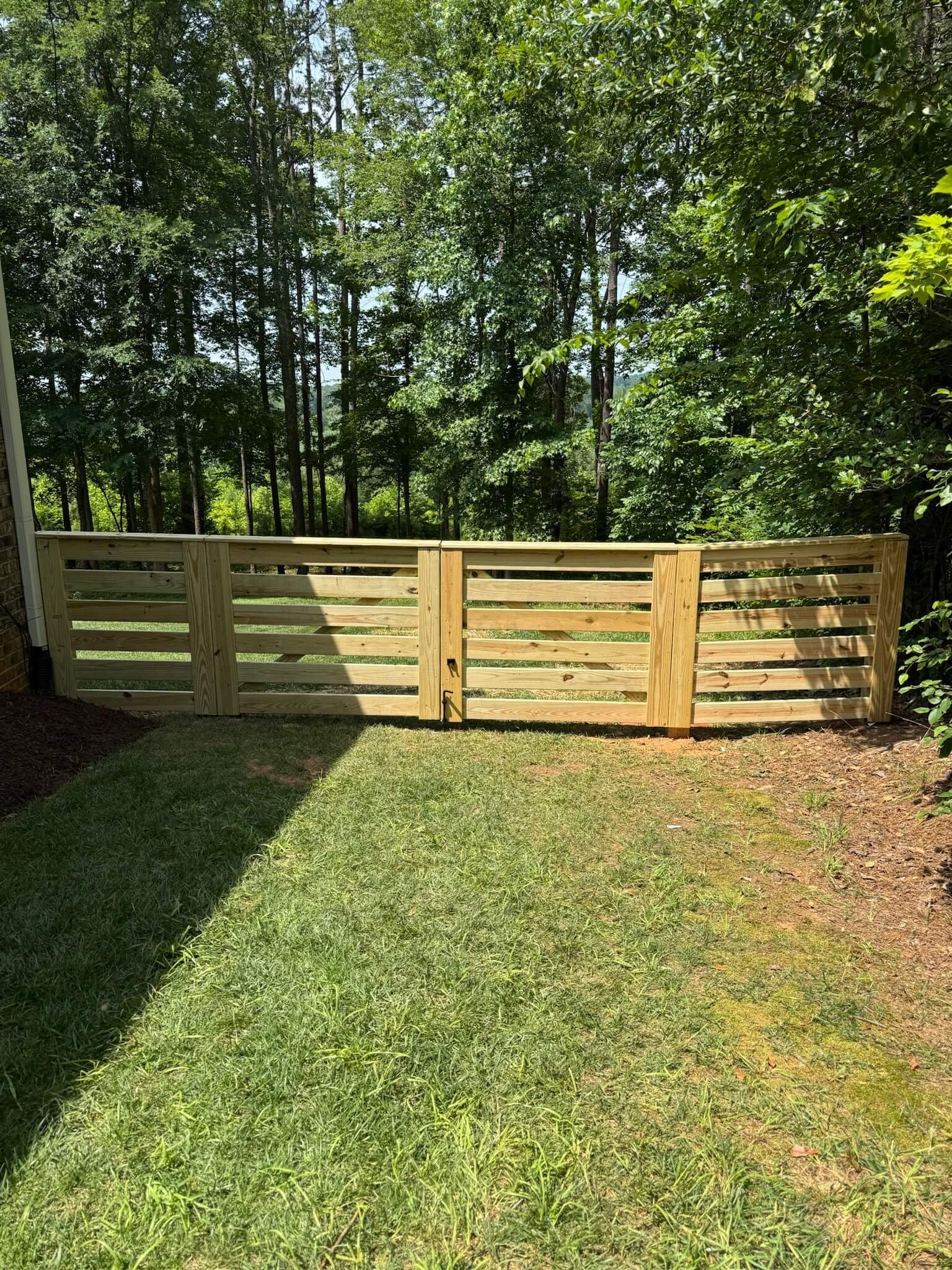 Sunlit backyard fence made of wooden slats beside a grassy lawn and trees