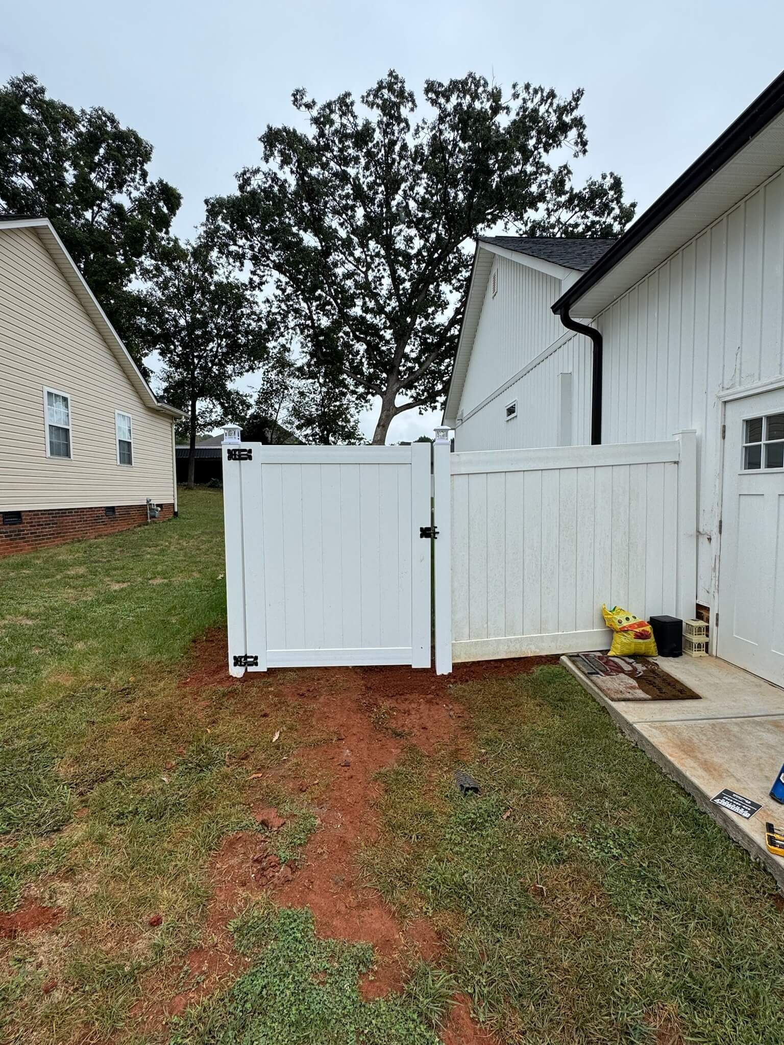 White gate between two houses on a sloped grassy yard, with red dirt and overcast sky.