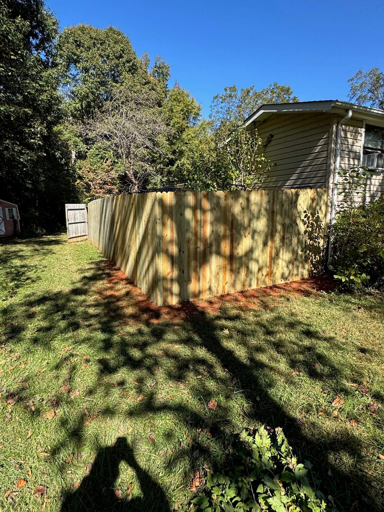 Backyard with wooden fence, house corner, trees, and grass in sunlight