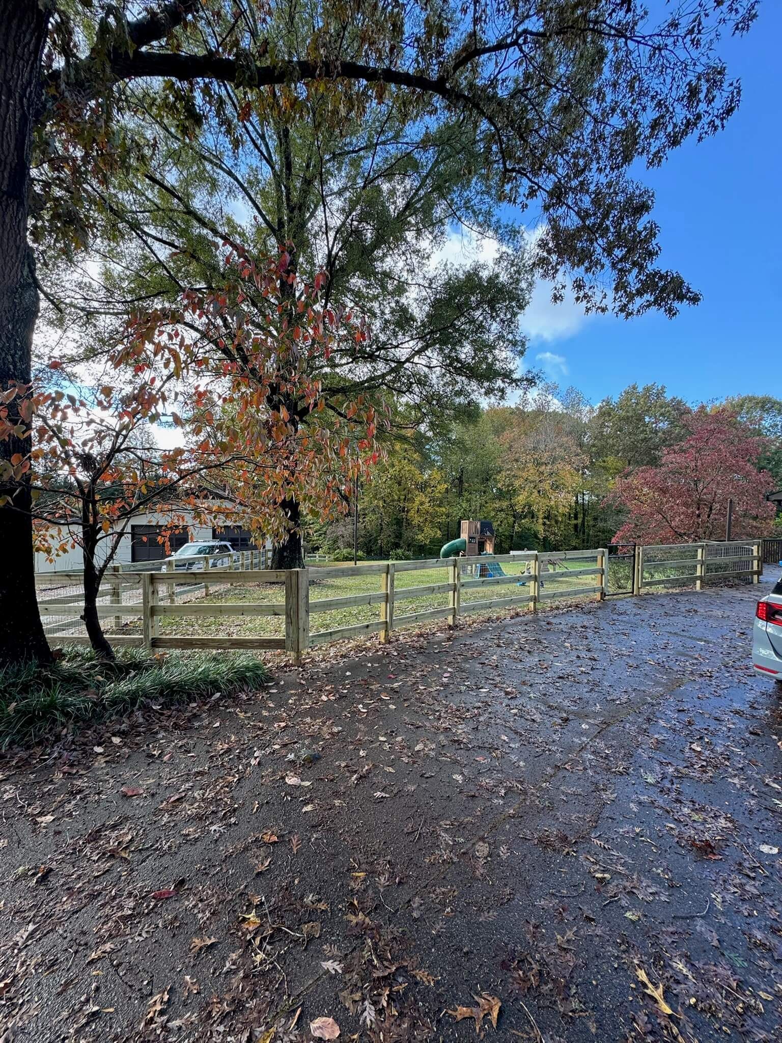 Autumn park path beside a wooden fence, with trees in fall color and a cloudy blue sky.