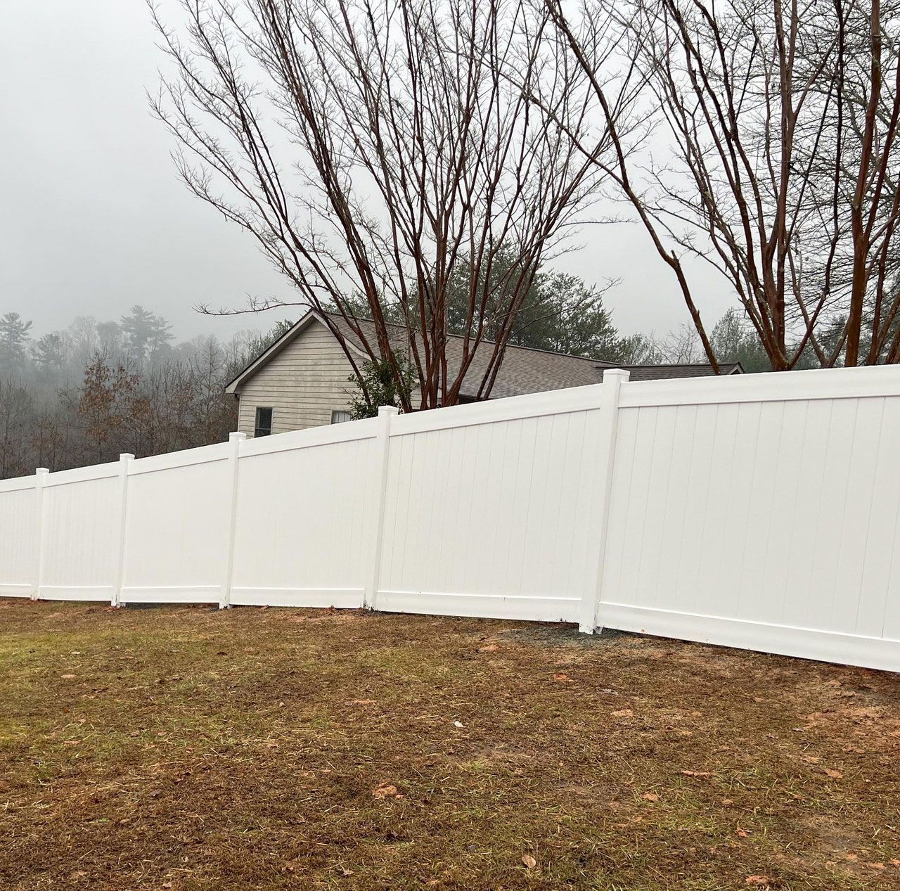 White vinyl fence in a yard with bare trees and a small house in the background under a gray sky