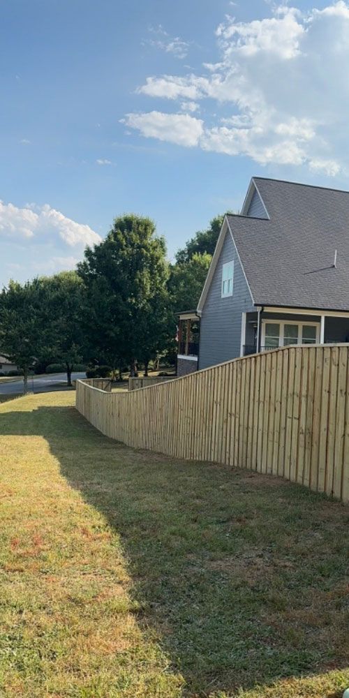 Suburban backyard with wooden fence, grassy lawn, trees, and a house under a blue sky with clouds