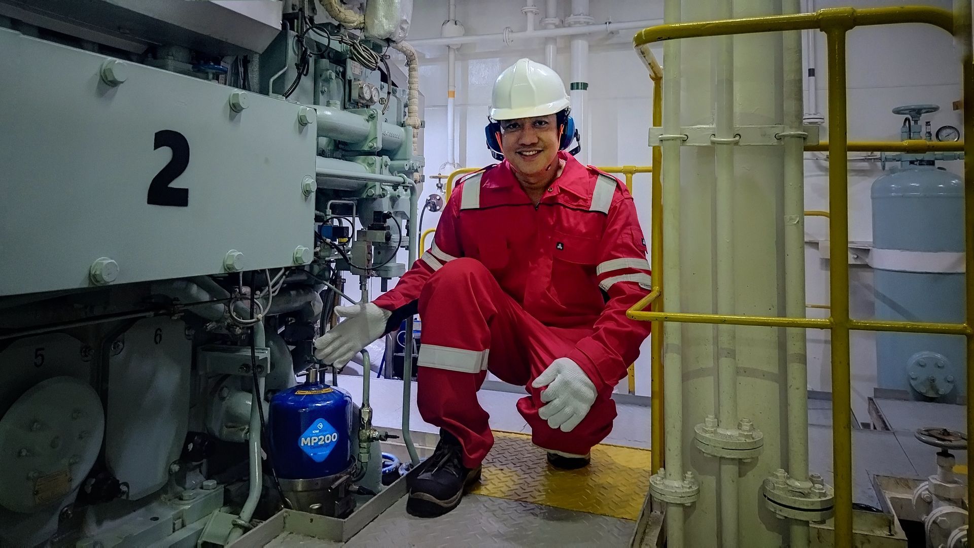 Engineer in red overalls and hard hat checks equipment in an engine room.