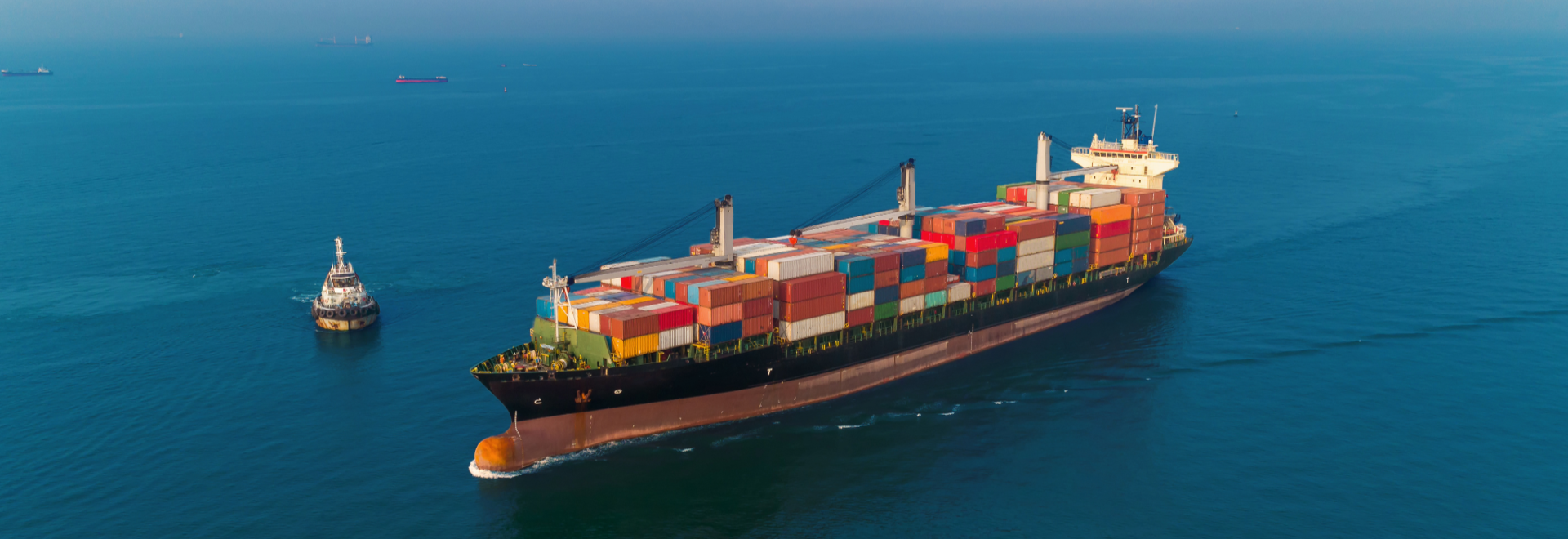 A container ship sailing on blue water, accompanied by a smaller boat. The ship is loaded with containers.