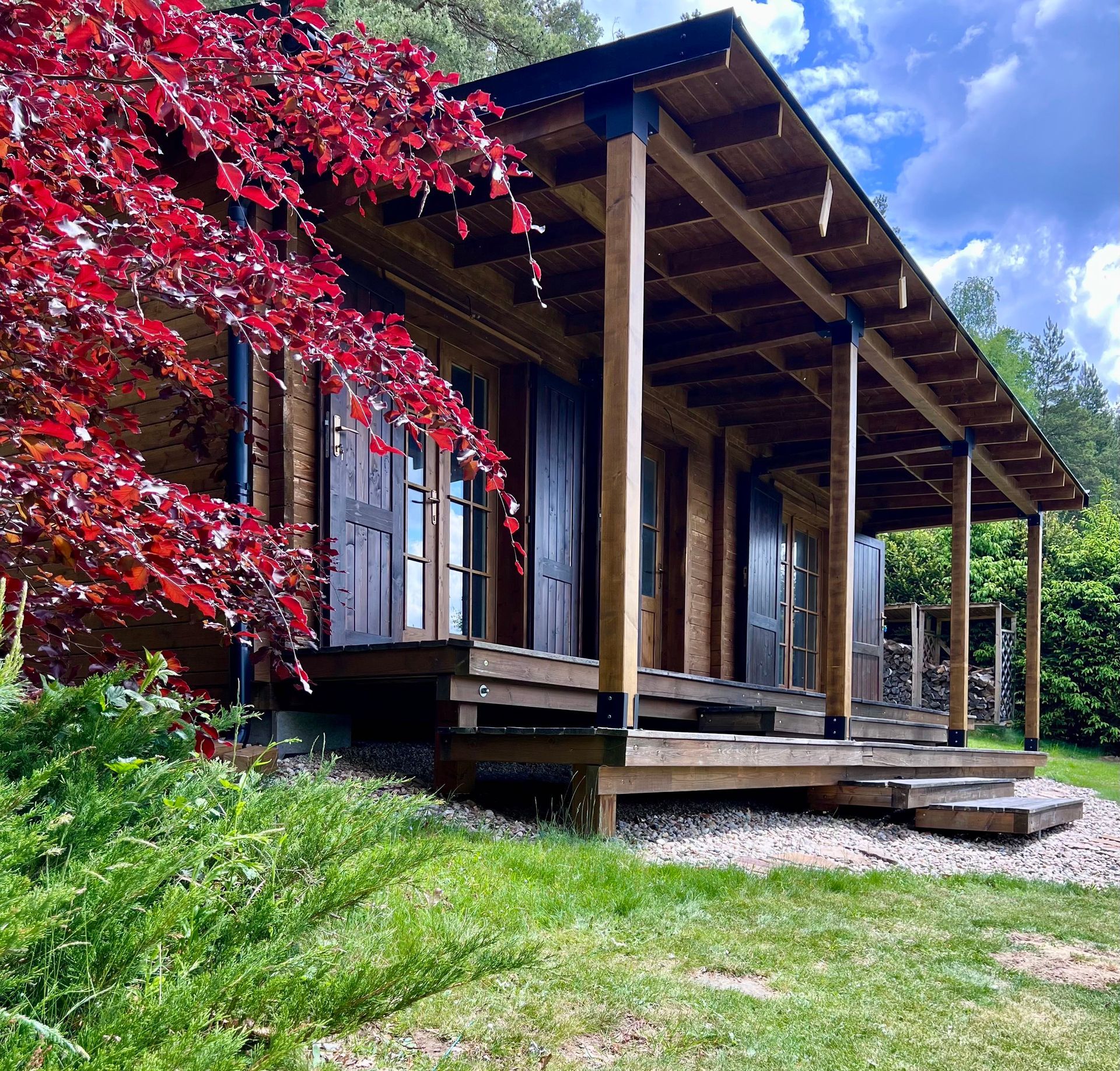 Wooden cabin with a porch, red-leafed tree in the foreground, on a grassy hill.