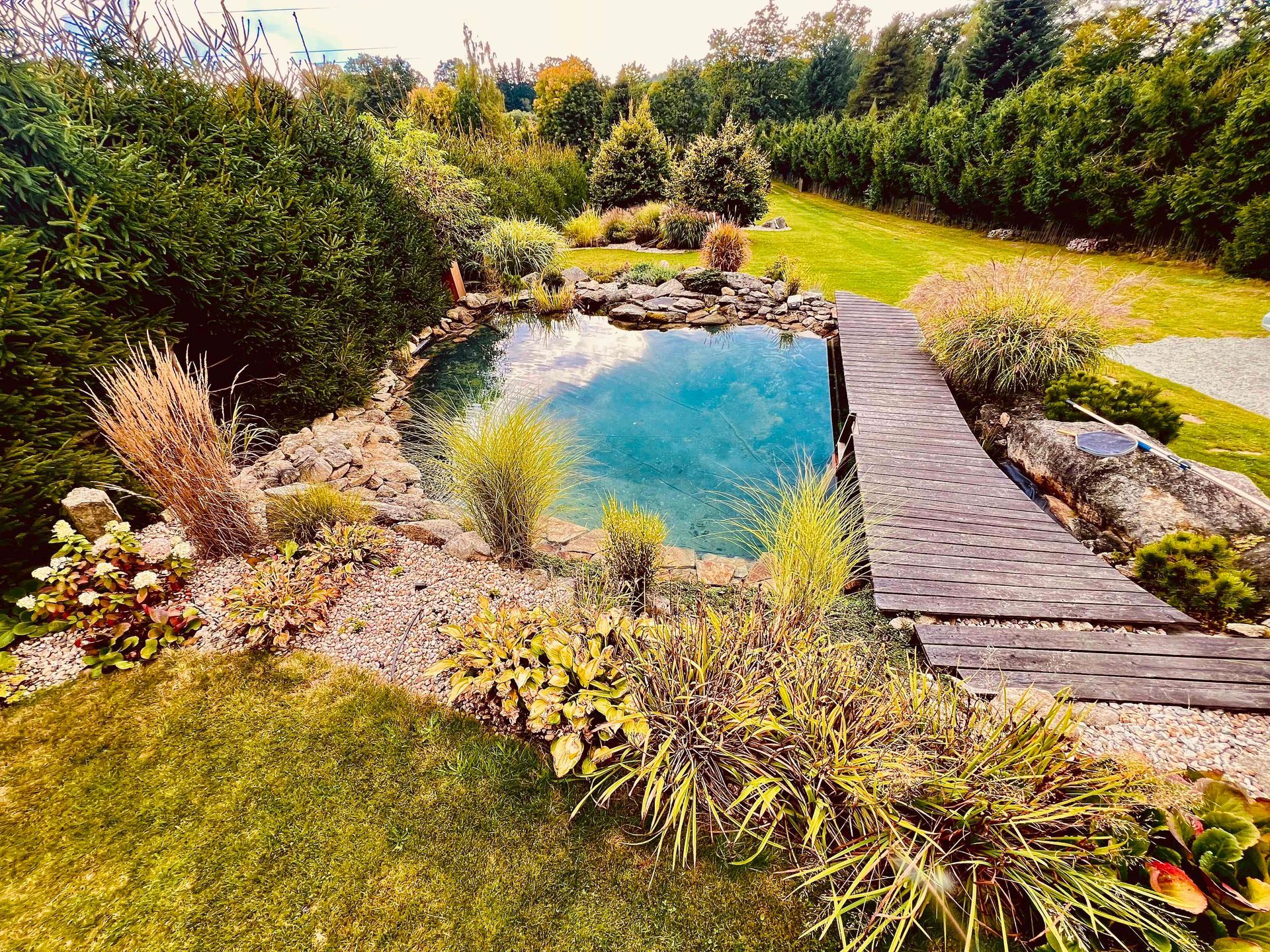 A serene garden with a pond, wooden path, lush plants, and a green lawn.
