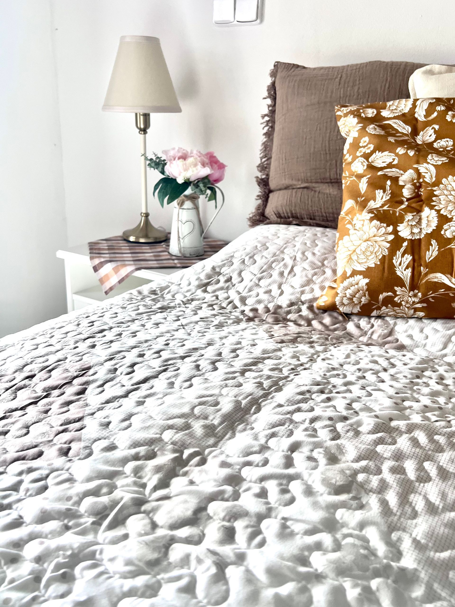 Bed with white textured quilt, side table with lamp and flowers, brown pillows.