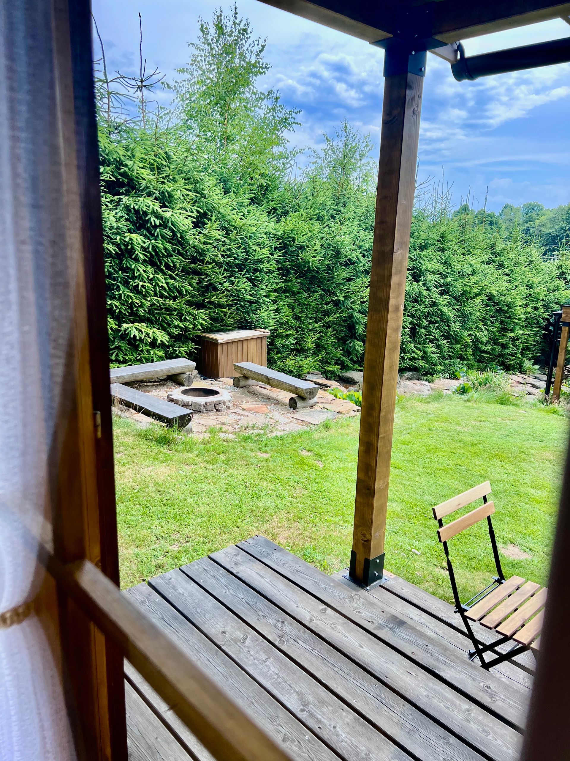 View from a cabin: wooden deck with chair, fire pit in green yard, lush trees in the background.