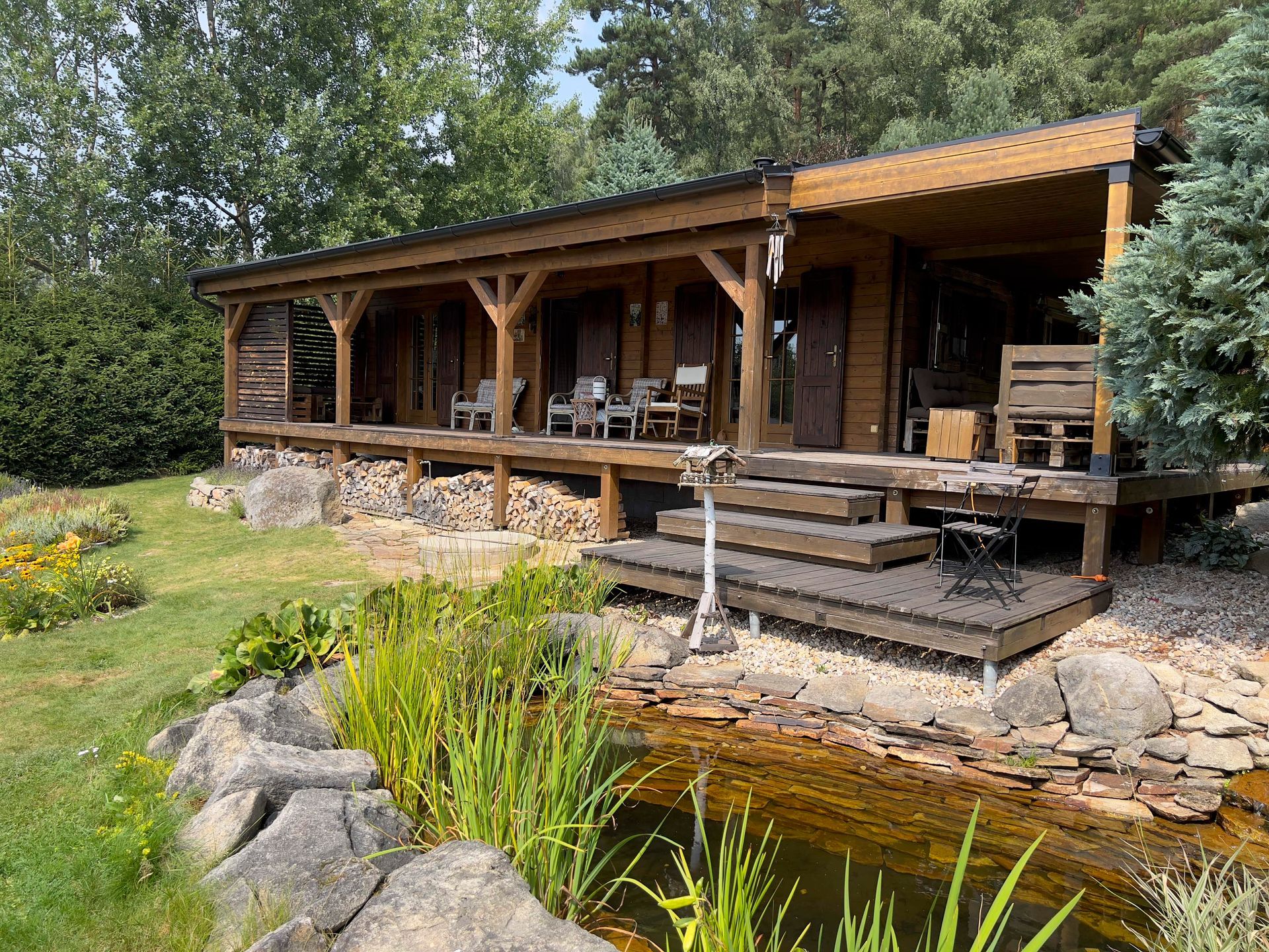 Wooden cabin with a porch overlooking a pond and garden; sunny day.