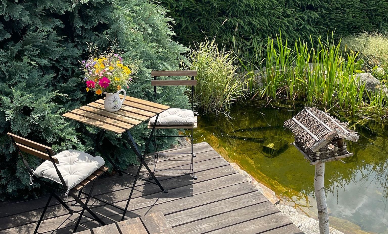A small table and chairs with cushions on a wooden deck beside a pond, birdhouse, and greenery.