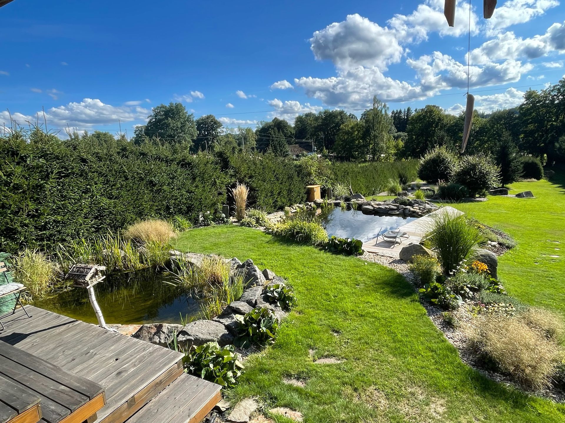 Lush garden with pond, green grass, shrubs, and a blue sky with clouds.