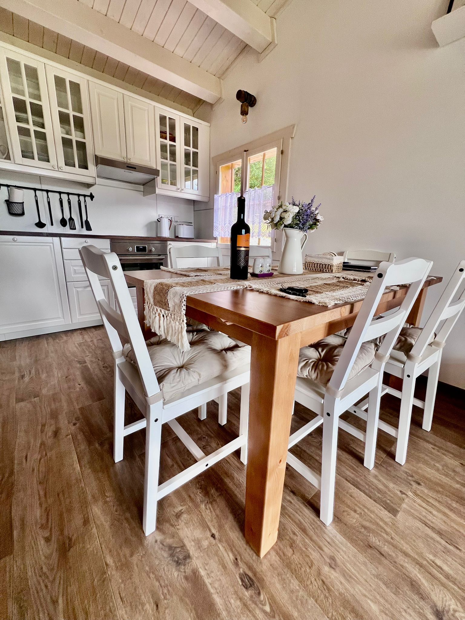 Wooden dining table with four white chairs, small kitchen with white cabinets and a window with flowers.
