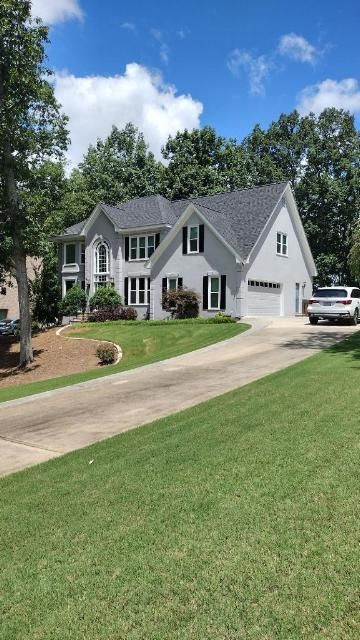 A light grey two-story house with dark shutters and a shingled roof, set back from a concrete driveway on a sunny day.
