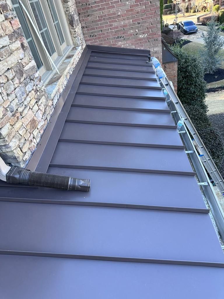 A dark brown, standing seam metal roof section installed beneath a brick wall and window, with a gutter along the edge.