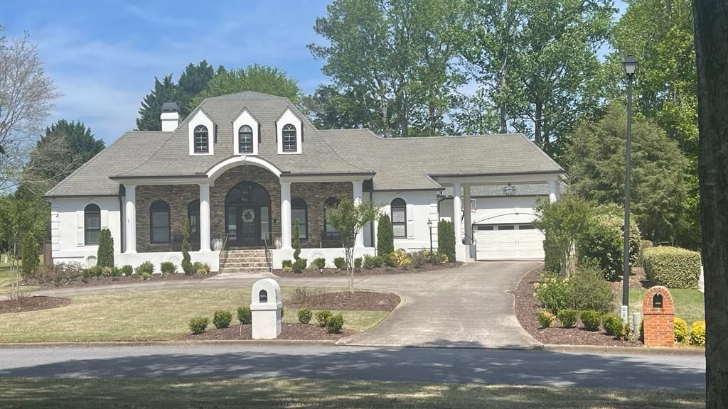 A white brick house with a stone entryway, three dormer windows, and a side-entry garage, set in a sunny residential yard.